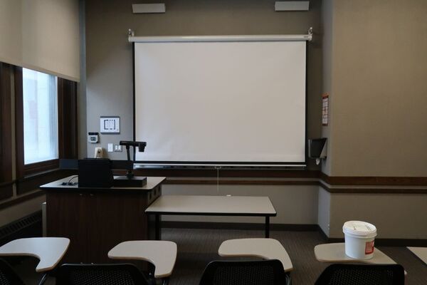 Front of room view with lectern on left in front of markerboard and projection screen lowered