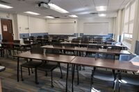 Back of room view of student table and chair seating, exit door on left wall, and markerboards on rear wall of room