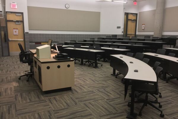 Back of room view of student tiered fixed-table and chair seating and exit door at left rear and front of room