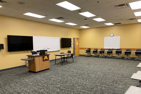 Front of room view with lectern on left in front of markerboard and display monitors on the left and right 