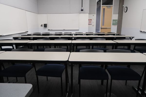 Back of room view of student table and chair seating, markerboards on left and rear wall, and exit door at right rear of room