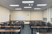 Back of room view of student table and chair seating, multiple markerboards on left side and back wall, exit door in left rear corner