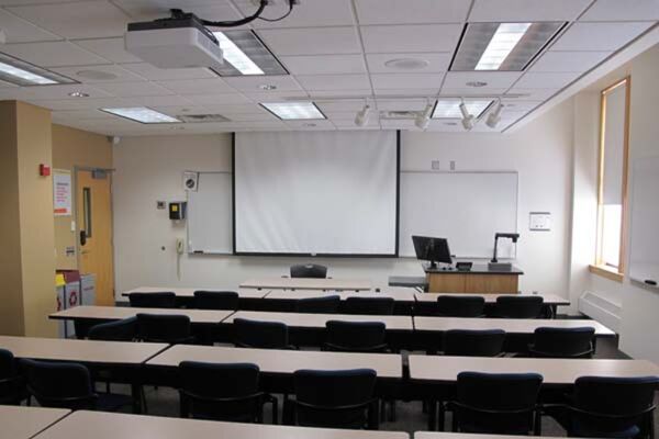 Front of room view with lectern on right in front of markerboard and projection screen lowered, exit door at front left