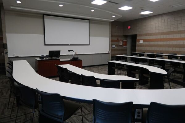 Front of room view with lectern on left in front of markerboard and projection screen lowered, exit door on the right