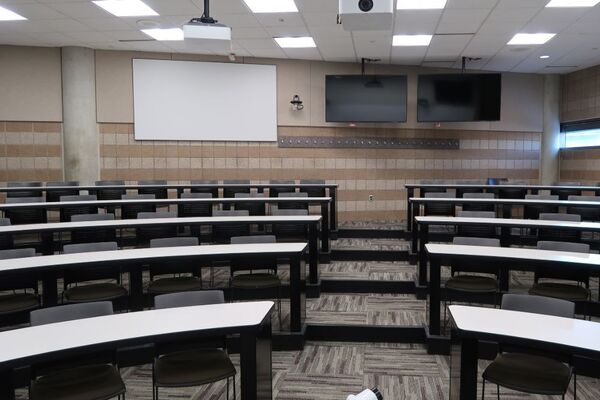 Back of room view of student tiered fixed-table and chair seating and projection screen center, confidence monitor on the right